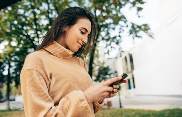 Beautiful stylish woman holding mobile phone outdoor on spring day. Pretty brunette female standing in the city street browsing online on modern smartphone. Woman watching social video on cellphone - Foto, Imagen