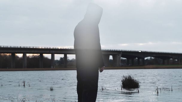 A bleak, moody, winter edit of a ghostly transparent hooded figure vanishing and standing next to a lake, looking out to a motorway bridge, with an out of focus in the background. - Foto, immagini