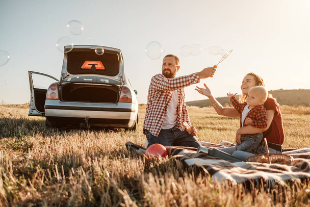 Feliz familia joven mamá y papá con su pequeño hijo disfrutando de un picnic de fin de semana de verano en el coche fuera de la ciudad, jugando con burbujas
 - Foto, imagen
