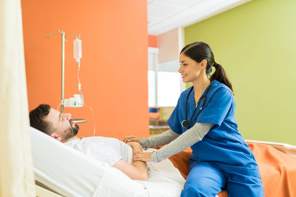 Uśmiechnięta młoda pielęgniarka pocieszająca mężczyznę podczas siedzenia na łóżku w szpitalu - Zdjęcie, obraz