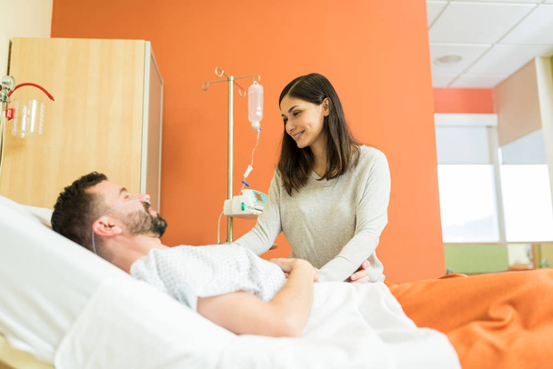 Smiling woman consoling male patient while standing by bed during hospital visit - Photo, Image
