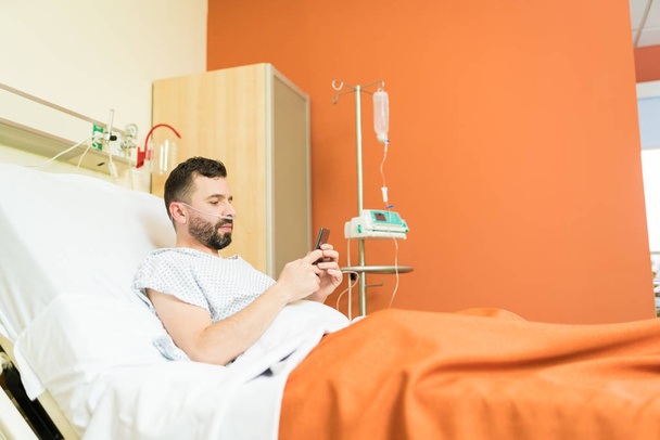 Hastanedeki yatakta dinlenirken akıllı telefondan mesajlaşan yetişkin bir erkek hasta. - Fotoğraf, Görsel