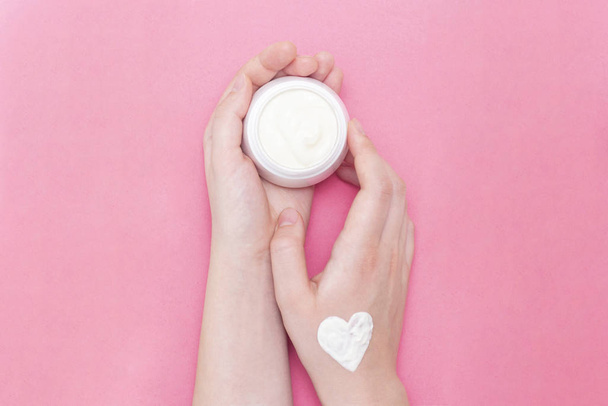 Hands of a beautiful well-groomed woman with a cream jar on a pink textural background. Moisturizer for clean and soft skin in the winter. The heart shape is made of cream. I love the body. Healthcare concept. - Fotografie, Obrázek