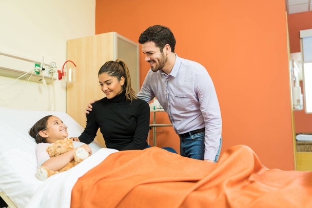 Hymyilevät vanhemmat katsomassa tytärtä, joka saa hoitoa sairaalassa käynnin aikana
 - Valokuva, kuva