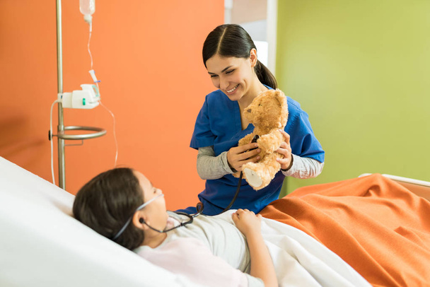 Молода медсестра і дівчина грають, тримаючи плюшевого ведмедя і стетоскоп в лікарні
 - Фото, зображення