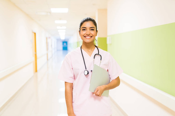 Usmívající se krásný hispánský lékař se stetoskopem a podložkou stojící v nemocniční chodbě - Fotografie, Obrázek