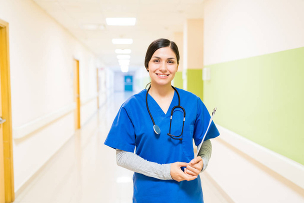 Latin young female doctor with stethoscope and clipboard wearing shrubs standing in corridor at hospital - Foto, imagen