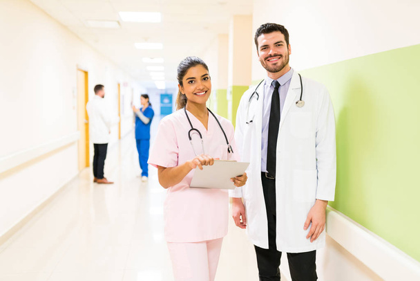Glimlachende Spaanse gezondheidswerkers staan aan de muur met collega 's op de achtergrond in het ziekenhuis - Foto, afbeelding