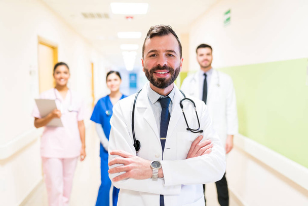 Latino medico medio adulto con le braccia incrociate contro i colleghi in piedi in background in ospedale
 - Foto, immagini