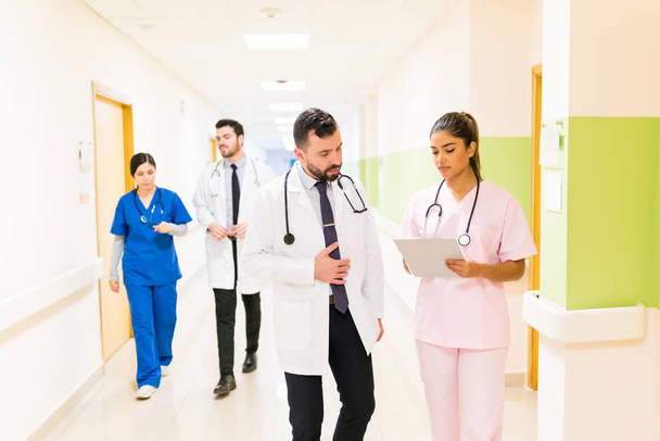 Sağlık çalışanları raporlar üzerinde tartışırken tıp arkadaşları hastane koridorunda arka planda yürüyorlar. - Fotoğraf, Görsel