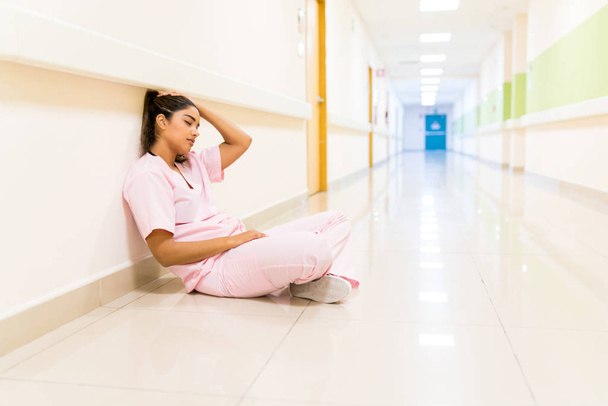 Uitgeputte jonge Latijnse verpleegster zit op de vloer tegen de muur in gang in het ziekenhuis - Foto, afbeelding