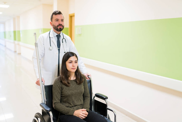 Средний взрослый врач с выпиской молодой женщины на инвалидной коляске в коридоре больницы
 - Фото, изображение
