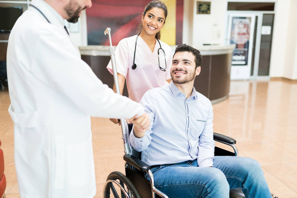 Hastane lobisinde doktorlarla el sıkışırken tekerlekli sandalyede gülümseyen bir erkek. - Fotoğraf, Görsel
