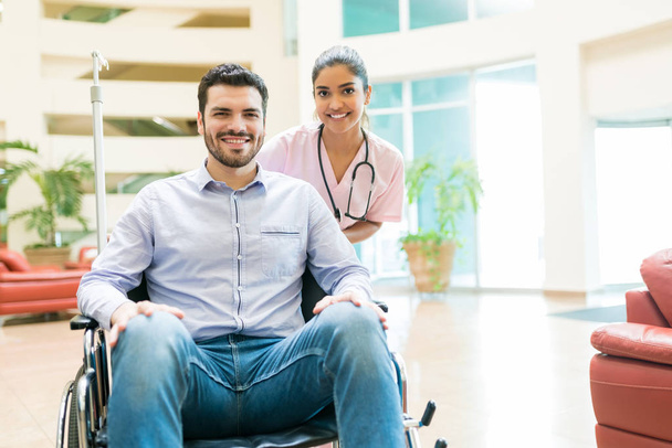 Sorridente inserviente latino con uomo medio adulto seduto sulla sedia a rotelle nella hall in ospedale
 - Foto, immagini