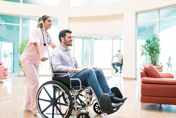 Junge Frau schubst Mann im Rollstuhl in Lobby des Krankenhauses - Foto, Bild