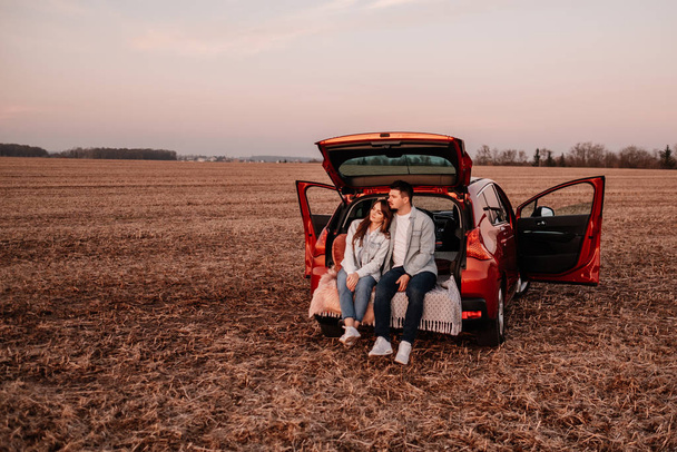 Jeune couple heureux habillé comme dans une chemise blanche et jeans assis à leur nouveau coffre de voiture, beau coucher de soleil sur le terrain, vacances et Voyage Concept
 - Photo, image
