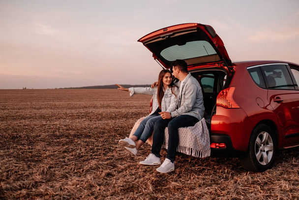 Молодые счастливые пара одеты как в белую рубашку и джинсы, сидя у своего нового автомобиля ствол, красивый закат на поле, отпуск и концепция путешествия
 - Фото, изображение