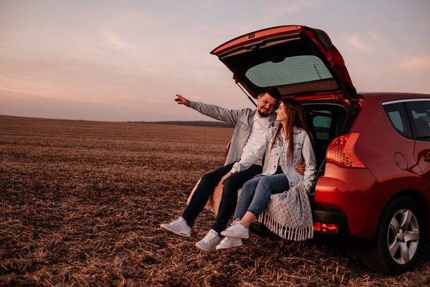 junges glückliches Paar in weißem Hemd und Jeans an seinem neuen Kofferraum sitzend, schöner Sonnenuntergang auf dem Feld, Urlaubs- und Reisekonzept - Foto, Bild