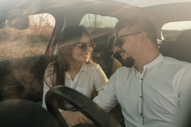 Giovane coppia felice vestita allo stesso modo in camicia bianca e jeans godendo di viaggio su strada alla loro nuova auto, bellissimo tramonto sul campo, vacanza e concetto di viaggio
 - Foto, immagini