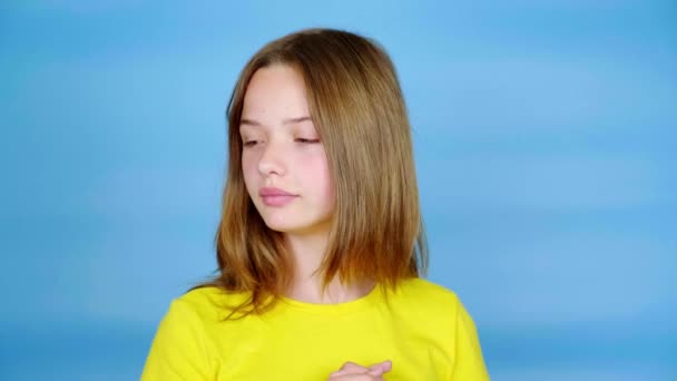 Tiener meisje in een geel t-shirt kijkt weg en beweegt haar lichaam in verschillende richtingen. Blauwe achtergrond met kopieerruimte. Tiener emoties. 4k-beelden - Video