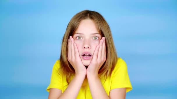 Teenie-Mädchen in gelbem T-Shirt ist sehr überrascht, legt Hände an den Kopf und öffnet den Mund. blauer Hintergrund mit Kopierraum. Teenager-Emotionen. 4k Filmmaterial - Filmmaterial, Video