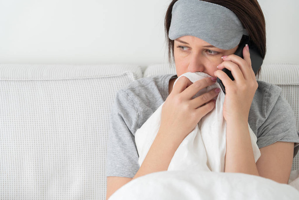 Сумна жінка носить спальну маску за допомогою смартфона, коли вона сидить у ліжку, покритому ковдрою
 - Фото, зображення