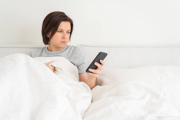 Kobieta za pomocą smartfona, jak siedzi w łóżku pokryte kołdrą - Zdjęcie, obraz