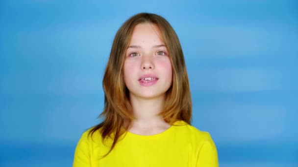 Sarı tişörtlü genç kız kameraya bakıyor başını sallıyor ve evet diyor. Kopyalama alanı olan mavi arkaplan. Gençlik duyguları. 4k görüntü - Video, Çekim