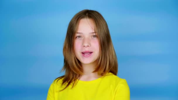 黄色のTシャツの10代の少女は、カメラを探している、彼女の頭をうなずき、はいと言う。コピースペース付きの青の背景。10代の感情4k映像 - 映像、動画