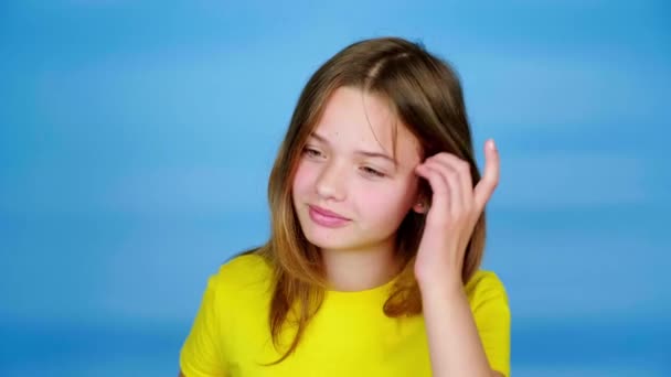 Teini-ikäinen tyttö keltaisessa t-paidassa suoristaa hiuksensa ja heittää ne takaisin. Sininen tausta ja kopiotila. Teini-ikäiset tunteet. 4k kuvamateriaali
 - Materiaali, video