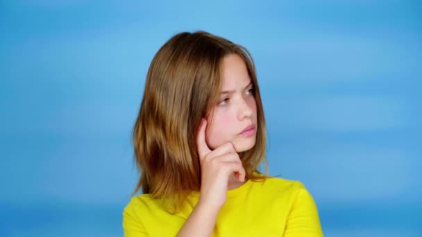 Tiener meisje in een geel t-shirt kijkt rond op de plaats voor tekst en denkt wat te kiezen. Blauwe achtergrond met kopieerruimte. Tiener emoties. 4k-beelden - Video