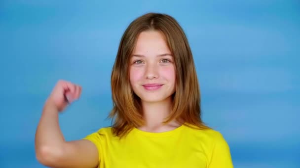 Η έφηβη με το κίτρινο μπλουζάκι δείχνει τους αντίχειρες πάνω και κοιτάζει την κάμερα. Μπλε φόντο με χώρο αντιγραφής. Εφηβικά συναισθήματα. 4k πλάνα - Πλάνα, βίντεο