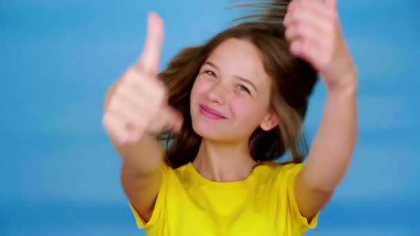 Happy teen girl in a yellow t-shirt is smiles, showing thumbs up, like and looking at camera. Blue background with copy space. Teenager emotions. 4k footage - Footage, Video