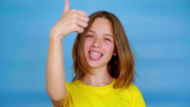 Chica adolescente feliz en una camiseta amarilla es muestra la lengua, pulgares hacia arriba, como y mirando a la cámara. Fondo azul con espacio de copia. Emociones adolescentes. Imágenes de 4k
 - Metraje, vídeo