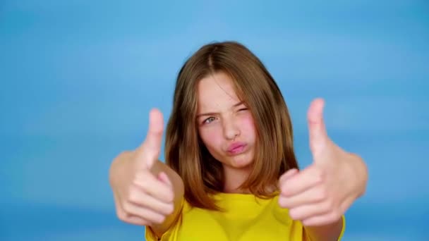 Happy teen girl in a yellow t-shirt is smiles, showing thumbs up, like and looking at camera. Blue background with copy space. Teenager emotions. 4k footage - Footage, Video