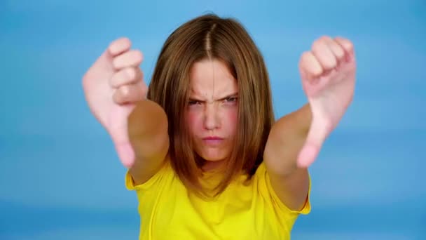 Sarı tişörtlü kızgın genç kız kameraya bakmaktan hoşlanmayan ve hoşlanmayan başparmaklarını gösteriyor. Kopyalama alanı olan mavi arkaplan. Gençlik duyguları. 4k görüntü - Video, Çekim