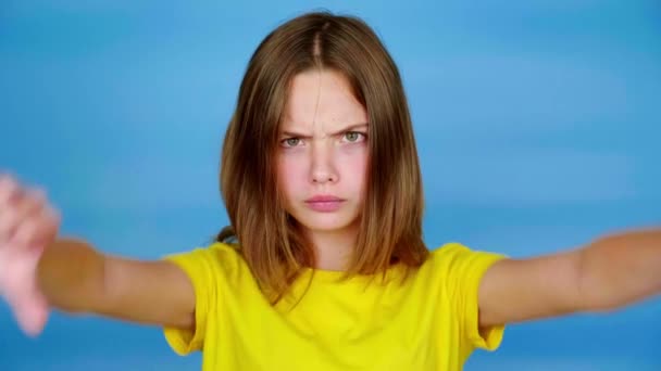 Menina adolescente irritada em uma camiseta amarela está mostrando polegares para baixo, não gosta e olhando para a câmera. Fundo azul com espaço de cópia. Emoções adolescentes. Filmagem 4k
 - Filmagem, Vídeo