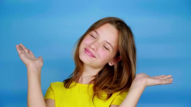 Joyeux adolescent fille dans un t-shirt jaune est ouvre son visage par les mains, souriant et posant, en regardant la caméra. Fond bleu avec espace de copie. Des émotions adolescentes. Images 4k
 - Séquence, vidéo