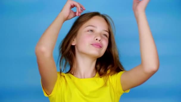 Happy teen girl in a yellow t-shirt is dancing, smiling and fools around. Blue background with copy space. Teenager emotions. 4k footage - Felvétel, videó