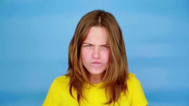 Tiener meisje in een geel t-shirt schreeuwt en kijkt naar de camera, grimmigen. Blauwe achtergrond met kopieerruimte. Tiener emoties. 4k-beelden - Video