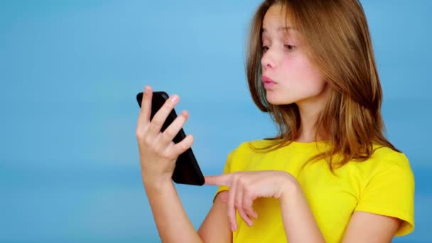 Η έφηβη με το κίτρινο μπλουζάκι είναι στο κινητό, έκπληκτη και λέει "ουάου". Μπλε φόντο με χώρο αντιγραφής. Εφηβικά συναισθήματα. 4k πλάνα - Πλάνα, βίντεο