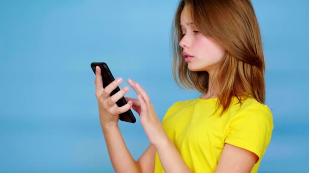 Teen girl in a yellow t-shirt is uses on smartphone, swipe touch screen. Blue background with copy space. Teenager emotions. 4k footage - Footage, Video