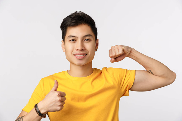 Czas to pompować. Atrakcyjny młody Azjata w żółtej koszulce pokazuje mięśnie i kciuk do góry, uśmiecha się, zachęca do ćwiczeń fizycznych, przychodzą na siłownię razem, chcą zdobyć silne bicepsy - Zdjęcie, obraz