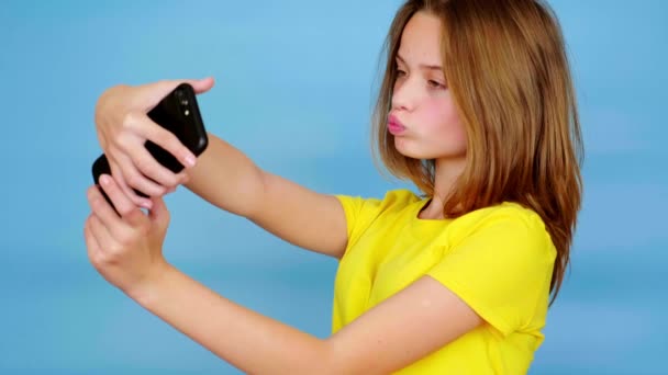 Ευτυχισμένος έφηβος κορίτσι σε ένα κίτρινο t-shirt είναι κάνει μια selfie ή βίντεο σε ένα smartphone και χαμογελώντας. Μπλε φόντο με χώρο αντιγραφής. Εφηβικά συναισθήματα. 4k πλάνα - Πλάνα, βίντεο