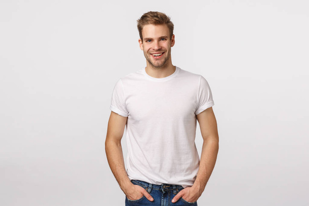 Charming, mature blond male model with bristle, wear white t-shirt, hands in pockets, smiling amused, look entertained, happy expression, agree with friend, standing enthusiastic white background - Foto, imagen