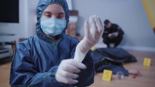 Portrait of forensic investigator in uniform putting on white gloves and looking at camera. Female professional working at the crime scene. Police officer sitting next to victim at the background. - Кадри, відео