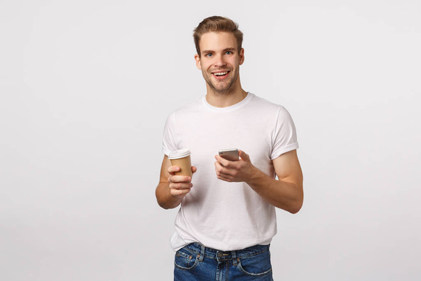 Cheerful good-looking modern blond man with bristle, holding paper cup of coffee, drinking tea and holding smartphone, messaging, smiling camera, browsing internet, shopping online during break - Foto, Bild