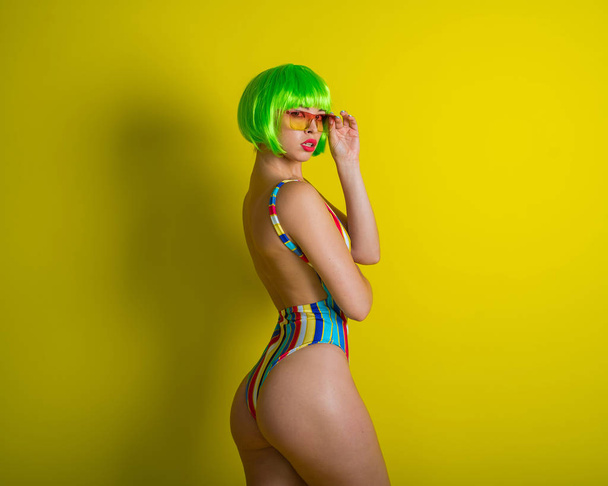Kaunis nainen vihreässä peruukissa ja raidallinen uimapuku poseeraa studiossa keltaisella taustalla. Tyttö kirkkaat hiukset ja punaiset huulet värillinen aurinkolasit
. - Valokuva, kuva