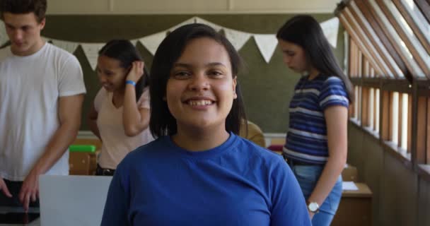 Portrait close up of a teenage mixed race girl with dark hair and brown eyes standing in a school classroom smiling to camera, with classmates talking in the background, slow motion - Felvétel, videó