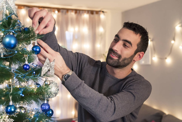 Mann mit Bart schmückt Weihnachtsbaum im Wohnzimmer. Urlaubsvorbereitung. - Foto, Bild